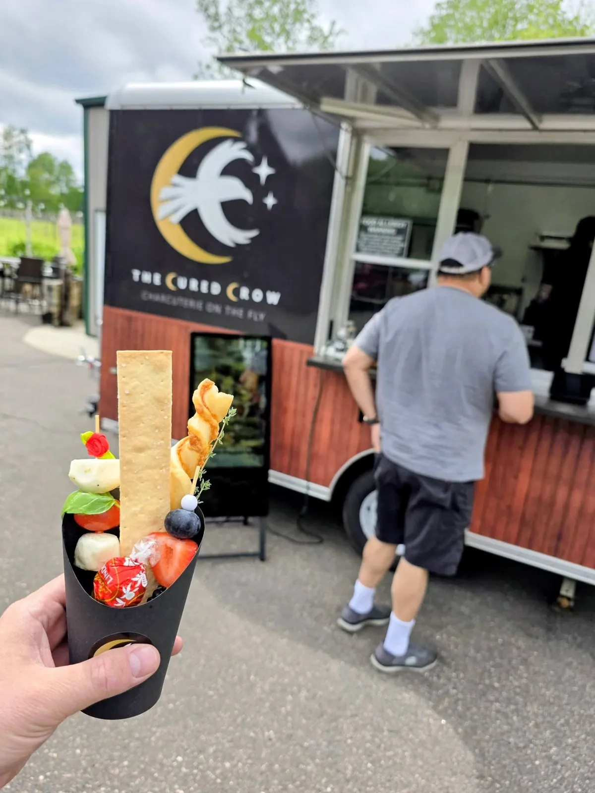 A Cured Crow charcuterie cup held in front of the trailer at a Champlin, Minnesota event