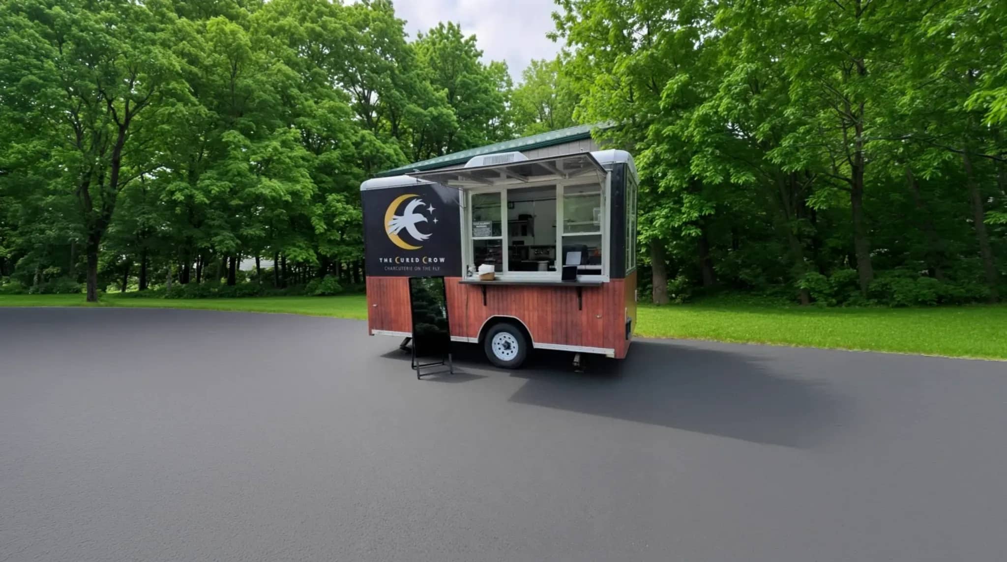 The Cured Crow branded charcuterie food trailer with its serving window open, parked against a backdrop of green trees